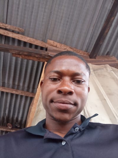 A selfie is taken from a low angle, focusing on the upper body of a young Black man in the foreground. He is wearing a dark blue collared shirt with two white buttons visible. His skin is dark brown, and he has short, dark hair. He is looking directly at the camera with a slight smile.
Behind him, the environment appears to be an outdoor or semi-outdoor setting with a corrugated metal roof supported by wooden beams and planks. The lighting suggests it is daytime. The background is partially obscured, with corrugated metal siding visible to the right.
The man's attire suggests a casual or work-related context. The overall scene conveys a simple, everyday moment captured through a personal photograph.
Based on the provided information, the image's location in Issele Uku, Nigeria, would indicate a domestic or semi-public space within a residential or semi-rural setting, characterized by the visible construction materials and roofing.
