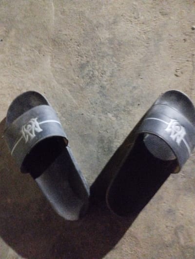 The scene, located in Kubare, Nigeria, depicts two dark grey synthetic slide sandals resting on a textured, light grey concrete surface. The sandals are positioned with their anterior sections diverging slightly, forming an approximate 'V' shape. Each sandal's upper strap features a white stylized "AM" monogram flanked by two thin horizontal white lines. Prominent, elongated dark shadows are cast posteriorly and inferiorly from each sandal, indicating illumination originating from the upper-left quadrant of the frame. No human subjects or active interactions are visible.