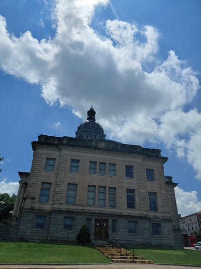 The image shows the exterior of a stone courthouse in Paris, United States of America. The courthouse features a central dome with a clock tower, topped by a spire. The building is constructed of light-colored stone blocks with darker stone at the base. There are multiple windows on each level of the building. In front of the courthouse, a manicured lawn slopes upwards towards the structure, and a set of concrete stairs with yellow-painted risers leads to the main entrance. A small, dark green tree is planted near the stairs. To the right of the courthouse, another building with a red bay door is visible, suggesting an adjacent commercial or municipal area. The sky above is blue with prominent white clouds, indicating a daytime scene. There are no people visible in the image.