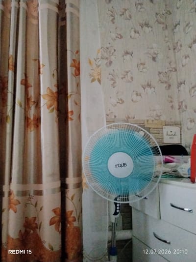 An indoor scene is captured, with vertical striped curtains featuring an orange floral pattern on the left side of the frame. To the right, a light blue oscillating fan with a white circular logo is prominently displayed. Behind the fan, a light-colored wall with a floral wallpaper pattern is visible. A white dresser with silver handles occupies the right side of the image, and on top of it are various items, including what appears to be paper and a red object. The bottom left corner of the image shows "REDMI 15," and the bottom right corner displays "12.07.2026 20:10." The location provided, "Айет, Kazakhstan," suggests this scene is within a residence or establishment in that region.