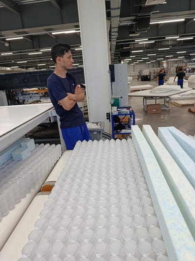 A man in a navy blue t-shirt and blue shorts stands with his arms crossed, facing left, in a factory setting. The background shows rows of white pocket springs, possibly for mattresses, and several long, rectangular pieces of blue and white foam. In the distance, other workers can be seen, along with factory machinery and shelving units stacked with materials. The overall environment suggests a manufacturing facility, specifically one involved in mattress production. This scene is located in the city of Zangiata, Uzbekistan.