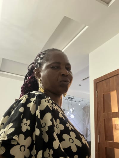 A woman with short hair and wearing gold earrings and a black and white floral print top is looking upwards. She is indoors, underneath a paneled wooden ceiling feature. Reflected in the ceiling is a chandelier. The location appears to be a building within the city of Mopo-Akinlade, Nigeria.