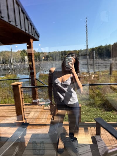 A female subject is depicted in reflection, taking a smartphone selfie through a large window while standing on a wooden deck in Otego, United States. The subject wears a light-patterned camisole and dark trousers, with her face partially obscured by the phone. The wooden deck features horizontal planking, a wooden post, and a cable railing. A small wooden table holding several magazines rests on the deck to the left. The foreground reflection on the glass shows a pair of dark binoculars and portions of two dark chairs. The background, visible through the glass, comprises a clear blue sky above a wetland containing numerous standing bare trees in water. Green vegetation borders the wetland, leading to a dense forest line in the distance. A structure with a light-colored roof is faintly discernible within the distant tree line. Part of the building's wooden eaves is visible in the upper left of the frame, indicating bright, clear daylight conditions.