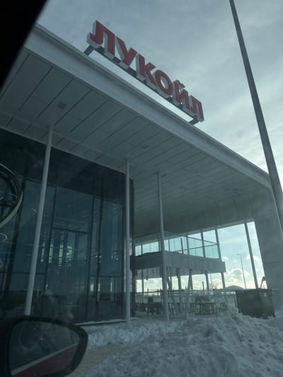 The image displays the exterior of a modern Lukoil facility in Vorsha, Russia, during winter. The ground in the foreground is covered by accumulated snow. The building features a light-colored facade with extensive glass panels and multiple white support columns. A prominent red sign displaying "ЛУКОЙЛ" (LUKOIL) in Cyrillic characters is mounted on the upper section of the building. An outdoor patio area, furnished with tables and chairs and enclosed by a glass railing, is visible beneath a protective overhang. Above this patio, an elevated, glass-enclosed structure is also present. The scene is captured from inside a vehicle, indicated by the partial view of a car's side mirror in the lower left. A tall, slender pole is positioned on the far right, and a dark container or dumpster is partially visible in the snow beyond the patio. The sky is overcast with patches of brighter light. No individuals are clearly discernible.