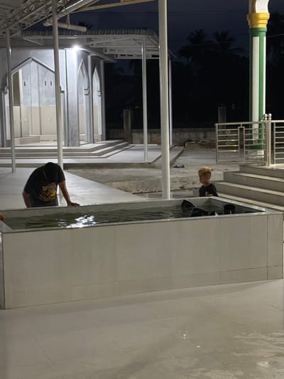 This image captures an illuminated ablution area within a mosque complex in the city of Bale, Indonesia, during nighttime. The scene is dominated by a rectangular, white-tiled water basin centrally located, containing water and several black ladles or scoops. An adult, visible from the torso up wearing a dark t-shirt, is leaning over the left side of this basin, with hands submerged or resting near the water's surface. To the right of the basin, a young child with distinctly blonde or light-colored hair and a dark t-shirt stands facing towards the basin or the adult.
In the background, a concrete structure featuring multiple arched openings and faucets, indicative of a wudu (ablution) station, is visible. This structure appears new or recently renovated, with clean lines and light gray finishes. White metal support columns and an overhead canopy extend across the scene. To the far right, a prominent architectural column with green and gold vertical stripes stands next to metal railings and a set of concrete steps. The ground is paved with light-colored tiles, exhibiting wet patches and reflections from the artificial lighting. Beyond the constructed area, the dark night sky and silhouettes of distant trees are discernible.