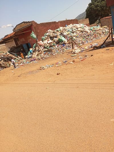 This image depicts a scene in Mukuyuni, Kenya, under a clear blue sky. The foreground features a reddish-brown dirt road, sparsely littered with small debris. To the left, a single-story building with a red corrugated roof and faded red walls displays "Airtel," "net," and "poa" branding above a blue door. Adjacent to this building, two small figures in orange and yellow attire are visible at ground level near the entrance. Further left, a person in a white shirt and dark trousers stands facing right. The central and right midground is dominated by a large, elevated pile of mixed refuse, predominantly plastic bags, bottles, and general waste, extending towards the background. This refuse partially obscures the building's rear. Wooden posts are visible at the refuse pile's right edge. Scattered debris from the main pile lies along the road's shoulder. Distant mountains and green trees are visible on the horizon, with utility wires crossing the upper left sky.