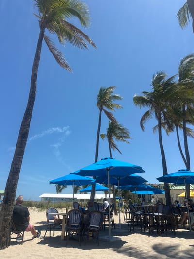 The scene shows a beachside cafe in Lauderdale-by-the-Sea, Florida. Blue umbrellas shade tables and chairs on the sand. People are seated at some tables, and the beach extends to the ocean in the background. Palm trees frame the scene against a clear blue sky.