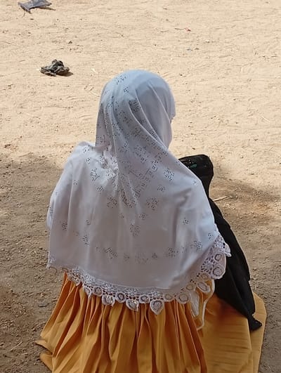 The image shows a person from behind, kneeling or sitting on dry, sandy ground in an outdoor setting in Fanisau, Nigeria. The individual is wearing a white hijab that features delicate perforated patterns and a decorative white crocheted lace trim along its lower edge. Beneath the hijab, a bright, pleated orange-yellow skirt is visible. A dark, possibly black, piece of fabric or garment is positioned on the ground next to the person's right side, under the edge of the hijab.
The ground is light brown and appears dusty, with a few scattered dark, unidentifiable objects in the background, possibly debris or small rocks. The bright lighting suggests it is daytime and likely sunny. No faces or specific emotional expressions are visible. There is no visible text in the frame.