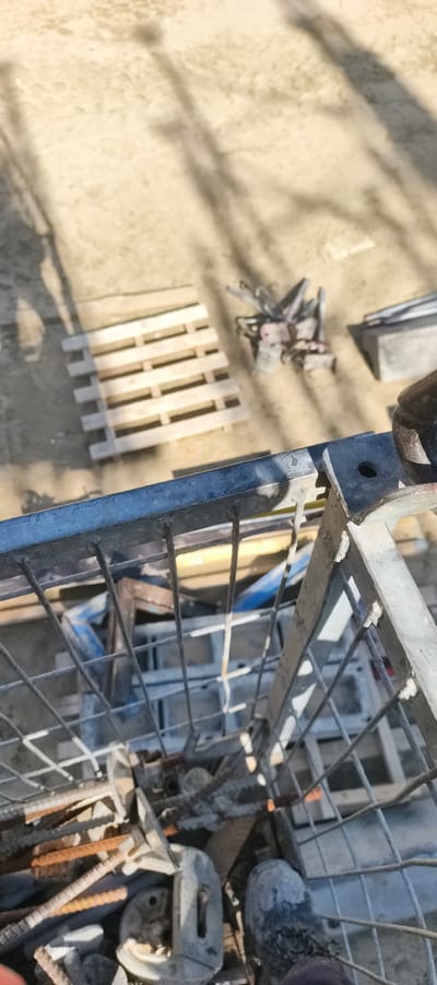 This image captures a construction or industrial scene, likely outdoors given the presence of dirt ground and shadows from sunlight. The foreground is dominated by a metal cage or bin filled with construction debris, including rebar, metal brackets, and other assorted hardware. A person's work boot, covered in dust or dirt, is visible at the bottom right of the frame, suggesting the photographer is standing within or beside this area.

In the mid-ground, a wooden pallet lies on the ground, alongside a pile of metal scaffolding components or similar equipment. Further back, a concrete block or formwork is visible. The overall setting appears to be a work area, possibly a construction site or a storage yard for building materials. The shadows cast on the ground indicate it is daytime, likely sunny.

The composition is shot from a high angle, looking down into the metal cage and out towards the ground. There is no visible text in the image. The scene is purely functional, conveying a sense of ongoing work and the materials involved. The focus is on the objects and the environment, with the human presence only implied by the boot. The context of Filino, Russia, adds a geographical location to this industrial setting.