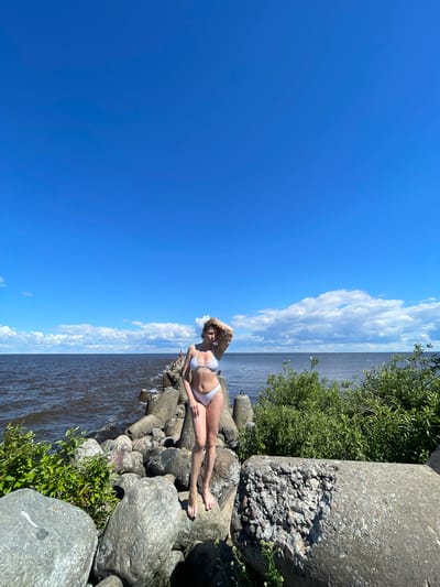 In a full shot taken outdoors, a woman stands on a rocky embankment, a body of water to her left, and a clear blue sky with sparse clouds above. She is positioned on a large grey boulder, wearing a white bikini, with her left hand touching her hair. The woman's pose is relaxed, with her weight shifted to her right leg. The embankment is made up of large, rough grey rocks and concrete structures. Green foliage is visible on the right side of the image, growing among the rocks. The water appears dark and slightly rippled. The overall impression is a sunny day in a coastal or lakeside environment, possibly a breakwater or pier. The location is the city of Belozersk, Russia, a historic city on the shores of Lake Beloye.