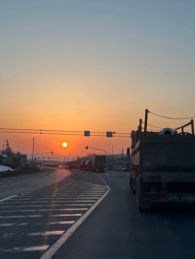 This image depicts a street scene in Shelepino, Russia, during either sunrise or sunset. The sky transitions from a light blue at the top to a bright orange-yellow gradient near the horizon, where the sun is visible as a distinct, intensely bright orange sphere.
In the foreground, a multi-lane asphalt road curves slightly to the left, featuring clear white dashed and solid lane markings, as well as a triangular hatched island on the left. The road surface exhibits reflective qualities, suggesting moisture. On the immediate right, the rear section of a dark-colored semi-trailer truck is prominently visible, featuring multiple rear wheels and chains strung across its upper frame. A partially visible rear license plate is present, consistent with Russian format.
Further along the road, in the mid-ground, a queue of vehicles is visible. This includes at least two more semi-trailer trucks (one appearing to be a tanker) and several smaller passenger cars, all heading away from the viewer. These vehicles appear stationary or moving slowly, indicating traffic congestion at an intersection ahead.
Overhead, a network of utility wires spans the roadway, from which multiple traffic lights are suspended; one light displays red. Several blue-and-white rectangular traffic signs are also affixed to the overhead structure. On the left side of the road, patches of snow or slush are visible along the shoulder, receding into a backdrop of sparse, bare trees and distant low-rise buildings. On the right side of the road, beyond the vehicles, industrial-style buildings are present. No discernible human figures are visible in the scene.