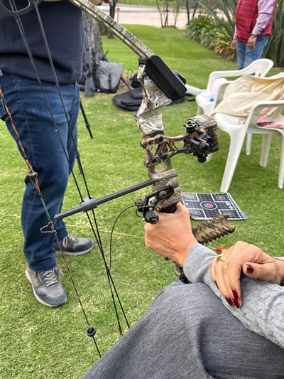 A compound bow is held by two individuals, one with jeans and grey sneakers, and the other wearing a grey sweater with red nail polish. The bow is camouflage, with a black sight and arrow rest. A target with concentric circles and grid lines sits nearby. The background features green grass, white plastic chairs, and other people. The bowstring is drawn back, ready for release.
