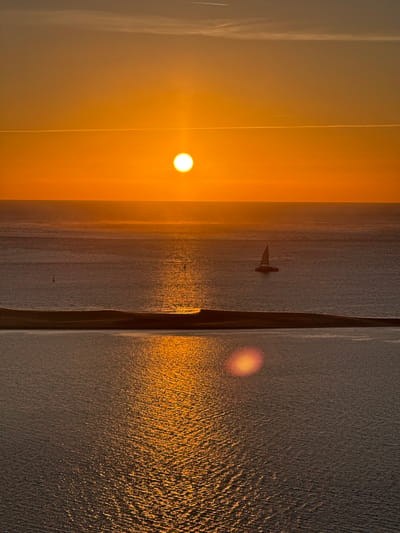 A wide-angle vertical shot captures a dramatic sunset over the ocean, likely near La Teste-de-Buch, France. The sun, a brilliant orange orb, hangs low in the sky, casting a warm glow and creating a shimmering reflection on the water's surface. The sky transitions from a deep orange near the horizon to a softer yellow and then a muted grey at the top. Two faint contrails from aircraft are visible in the sky.
In the middle ground, a narrow strip of land, possibly a sandbar or coastline, divides the ocean into two distinct bodies of water. The ocean itself is calm with gentle ripples. A lone sailboat with its sail up is visible on the further expanse of water, positioned to the right of the center. In the foreground, the water is a darker shade, reflecting the sunset colors more intensely, with a bright lens flare appearing as an orange circle on the right side. Two small buoys are faintly visible in the middle body of water. The overall atmosphere is serene and tranquil, characteristic of a coastal sunset.