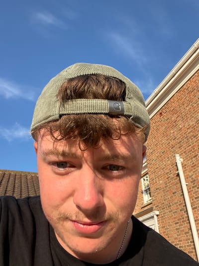 A person is facing the camera, slightly angled. They wear a corduroy cap, with curly hair peeking from underneath. Visible features include their eyes, nose, and lips. A beaded necklace is around their neck. The background consists of a clear blue sky, the top of a brick building, and a white pipe running along the building. The lighting is bright, casting shadows on the face and adding to the clarity of the sky. The overall composition suggests an outdoor setting on a sunny day.