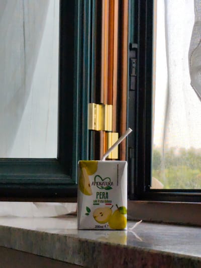 The image shows a close-up of a juice box on a windowsill, with a window in the background. The juice box, which is white and labeled "AMONATURA PERA," contains pear juice and has a straw sticking out. The window is dark-framed and has blinds or shutters behind it, also dark-framed. Through the window, a blurry green landscape is visible, suggesting an outdoor scene. The lighting suggests it is daytime, possibly late afternoon, given the soft, diffused light. The overall impression is one of domestic quietude, with the juice box suggesting a moment of refreshment or a simple snack. There are no people visible in the image. The setting appears to be indoors, near a window, likely in a residential or hospitality environment.