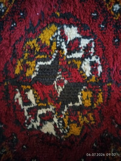 The image is a close-up, overhead shot of a patterned rug. The rug features a central medallion design with intricate geometric patterns in shades of deep red, black, white, and yellow. The texture appears to be that of thick pile, typical of a hand-knotted or machine-made rug. The lighting suggests an indoor environment, possibly artificial light. A timestamp in the bottom right corner reads "06.07.2026 09:02". The rug is not explicitly identified as being in Gurlan, Uzbekistan; this context is provided externally and cannot be verified from the image itself.