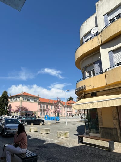 The image displays an urban square in Chaves, Portugal, under a clear blue sky with minimal white clouds. In the foreground, a female with dark hair, wearing a light pink long-sleeved top and white trousers, sits on a slatted wooden bench with dark metal supports, positioned in partial shadow on a cobblestone-like surface. Her right arm is raised towards her face.
To the left, several vehicles are parked parallel to a curb, including a silver Volkswagen Polo facing right and a black station wagon behind it. The central paved area of the square contains multiple rough-hewn grey concrete blocks and a large, blue, three-dimensional sign spelling "Chaves." Two tall, grey streetlights are also visible.
In the background, a large, multi-story building with a pink facade, numerous windows, and an orange tiled roof stands prominently, flanked by trees with pink blossoms. To the right, a curved, multi-story building with a yellow/beige facade and white sections occupies the frame. This building features balconies with blue and white patterned tile railings, white roll-down shutters on the windows, and a satellite dish mounted on an upper balcony. The ground floor displays a light-colored awning over a shop window, with a partially visible sign above reading "CAMISARIA S" and a street sign below identifying "Rua Candido Sotto Mayor."