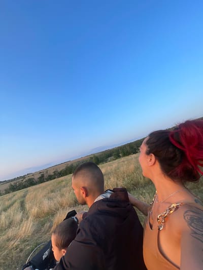 The image shows three people in a grassy field under a clear blue sky, likely during sunset due to the warm lighting. The perspective is tilted, giving a sense of being on an incline or looking down from a height.
In the foreground, a person with bright red hair tied up is leaning forward, their profile visible as they look down. They are wearing a beige top and a prominent gold chain necklace. Their right arm is bent, and they have tattoos on their upper arm.
Behind and below this person, another individual with a shaved head is also leaning forward. They are wearing a black jacket over a white shirt. Their head is tilted downwards, and they are looking towards something out of frame.
In the lower right of the image, partially visible and behind the person in the black jacket, is a third individual, also with a shaved head, looking downwards. They appear to be a child, possibly being held or embraced by the person in the black jacket.
The setting is an open, dry grassy field with some scattered trees and a distant, hazy landscape. The vast expanse of the blue sky dominates the left side of the frame. The overall mood suggests a moment of shared observation or connection between the individuals, possibly a family or friends enjoying the outdoors at dusk. No visible text is present in the image.