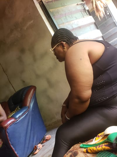 A middle-aged, dark-skinned woman with braided hair is wearing glasses, a black top, and black leggings. She has a silver bracelet on her wrist. She is indoors, near a window with what appears to be corn stalks on the outside ledge. She is seated next to a blue and maroon chair and orange sandals. The location is Umundugba, Nigeria.