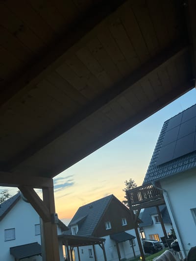 This image depicts an outdoor scene, likely a residential area, at dusk. The perspective is from beneath a wooden overhang, looking up towards the sky and neighboring houses. The sky displays a gradient of colors, transitioning from a pale blue to warm oranges and yellows, indicating either sunrise or sunset.
In the foreground, the dark, wooden structure of the overhang dominates the lower left of the frame, with visible wooden planks and structural beams. To the right and further back, several houses with dark tiled roofs and white facades are visible. Some houses have windows, with lights on in a few, suggesting indoor activity. One house features a balcony with a wooden railing. Solar panels are installed on the roof of one house.
A small white object, possibly a security camera or sensor, is mounted on a horizontal wooden beam in the mid-ground. A tree is partially visible behind one of the houses. The overall atmosphere is tranquil, enhanced by the soft evening light and the residential setting. No people are clearly visible. The image appears to be taken in Milná, Czechia, based on the provided location context.