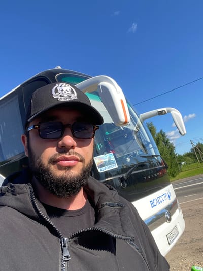 In the city of Kaykino, Russia, a man with a beard and sunglasses is taking a selfie in front of a white and black bus under a clear blue sky. The man is wearing a black baseball cap with a skull logo and a black hooded jacket. The bus has the text "ВЕЛЕСТР" and a license plate "E936YK 762" visible on its rear. A sticker that says "АЭРОПОРТ" is visible on the bus's windshield. The background shows a rural landscape with trees and a road.