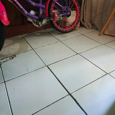 The image shows a white tiled floor in a home or building in Pameungpeuk, Indonesia, located at -7.641 latitude and 107.728 longitude. The floor is predominantly white with light gray grout. The tiles are square and are arranged in a grid pattern. The tiles are slightly dirty, with visible dirt smudges. There is a bicycle tire visible in the upper left corner of the image. The image is taken from a low angle. The focus is on the floor, with the bicycle tire being out of focus and only partially visible.  The scene suggests a quiet, domestic setting, potentially in a garage or storage space.