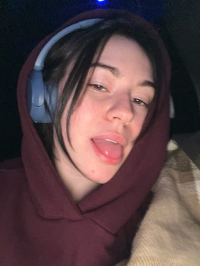 A person in a burgundy hoodie and gray headphones tilts their head, sticking out their tongue. Dark hair frames their face. The background includes a dark area and a patterned beige surface, possibly a blanket or cushion. The overall lighting is dim, creating a casual, informal mood.