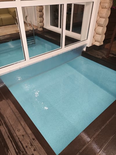 This media file captures an indoor swimming pool area with a rustic, cabin-like aesthetic. The pool itself is filled with clear, light blue water, appearing inviting and serene. The bottom of the pool has a speckled texture. A stainless steel ladder with a curved handle is partially submerged, indicating the pool is ready for use.

The pool is surrounded by a wooden deck made of dark brown planks, some of which appear wet, suggesting recent use or the humid environment of a pool area. To the left of the pool, large glass windows with white frames offer a view into another part of the building, where a light-colored sofa and a tiled floor are visible.

The structure of the building is notable, with a prominent wall constructed from rounded logs, characteristic of a log cabin. This log wall, with its hexagonal patterns where the logs meet, is a strong visual cue to the location's style. White window frames and metal accents are visible.

The lighting suggests it could be evening or an overcast day, as there are no strong shadows indicating direct sunlight. The overall atmosphere is peaceful and relaxing, hinting at a private retreat or a spa-like setting. There is no visible text in the image. The context provided, Старое Мартьяново, Russia, reinforces the impression of a cozy, potentially rural or resort-like environment.