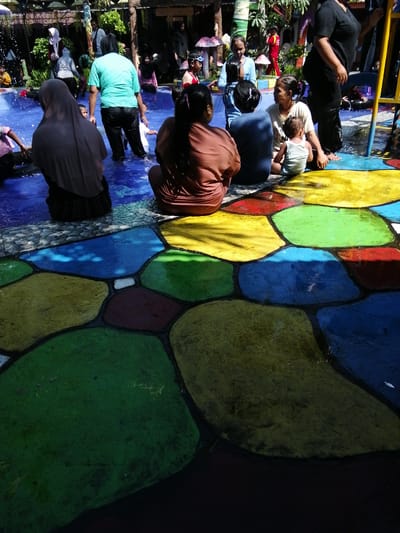 The image depicts a vibrant, colorful scene at a public swimming pool, likely located in Tegalwangi, Indonesia. The ground around the pool is a mosaic of large, irregular shapes painted in bright blues, greens, yellows, and reds, creating a playful and inviting atmosphere. Several people are visible, mostly dressed in swimwear or casual clothing, enjoying the water and surrounding area. In the foreground, a woman wearing a headscarf and a modest outfit is seated, facing away from the camera. Adjacent to her, another woman in a brown top is also seated. Further back, a group of individuals, including women and children, are gathered near the edge of the pool. Some are wading in the water, while others are standing or sitting. The presence of lush greenery and wooden structures in the background suggests an outdoor recreational setting. The overall impression is one of a lively community gathering at a water park or public pool, with a focus on family-friendly activities.
