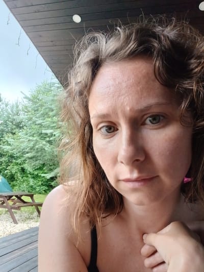 A close-up, selfie-style photograph shows a woman with light skin and medium brown, wavy hair. Her face is turned slightly towards the camera, and she has light green eyes and light pink lips. She is wearing a black top. In the background, there is a wooden deck with a picnic table and trees visible. The sky is overcast. The image appears to have been taken outdoors in a residential area, possibly in Verkhneye Dubrovo, Russia.