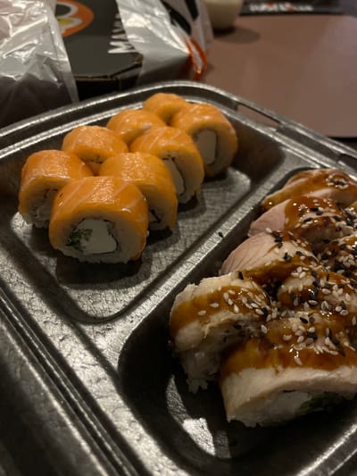 A top-down, close-up shot shows a grey disposable food container divided into two sections. The container is filled with sushi rolls. In the left section, salmon-covered rolls are visible, revealing white cream cheese and small pieces of green filler inside. In the right section, white-meat rolls are drizzled with a brown sauce and sprinkled with white and black sesame seeds. The left part of the image shows a small part of packaging. The photograph does not indicate the location of Sepych, Russia.