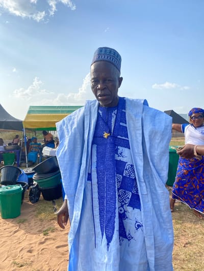 An elderly man, appearing to be African, stands in the foreground, facing the camera with a direct gaze. He wears a light blue traditional garment with an intricately patterned darker blue inner tunic and a matching blue cap. A gold-colored pendant is visible on his chest. To his left, a yellow and green striped canopy shades several seated individuals and tables with bowls and containers. Below, a stack of black, green, and grey plastic buckets and basins rests on the sandy ground. To the man's right, a woman, also appearing African, is partially visible. She wears a white top with a blue logo, a patterned blue and orange skirt, and a blue headwrap, with her right arm extended horizontally. In the far background, a black canopy is visible. The scene is outdoors under a blue sky with scattered white clouds, on sandy terrain in Itobe, Nigeria.