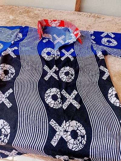 An overhead view captures a long-sleeved garment, likely a shirt or tunic, laid flat on a light beige, textured surface. The garment features a multi-paneled design. The central body is dark blue or black, adorned with large, abstract white "X" and "O" motifs interspersed with vertical, wavy white and thin dark blue stripes. The collar is patterned in red and white, displaying an abstract, organic design. The upper shoulder and sleeve sections are a royal blue with white horizontal stripes and fragmented white "O" shapes. The fabric appears to be cotton or a similar textile. The visible environment is an indoor setting, with the textured surface possibly being a bedspread, tablecloth, or similar fabric-covered surface. No individuals are present, and no actions or interactions are depicted.