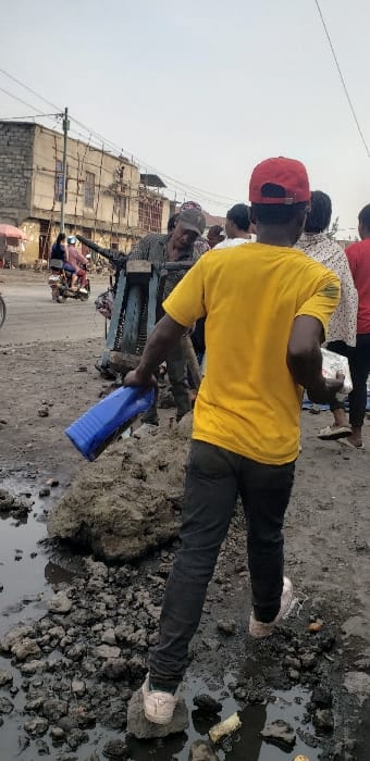 The image captures an outdoor urban scene in Buzove, Democratic Republic of the Congo, under an overcast sky. The foreground features an uneven, muddy dirt ground with standing water in puddles and large clumps of dark soil. A person, viewed from behind, is centrally positioned, wearing a red baseball cap, a plain yellow t-shirt, dark trousers, and light-colored sneakers. Their left foot is placed on a large lump of mud. The person's right hand holds a blue plastic container, possibly a bottle or jerry can, with a silver or metallic band near its base.

In the midground, to the left, a person wearing a plaid shirt and a dark cap leans towards a large, blue-grey metallic structure, which appears to be a piece of equipment or a heavy cart with a visible black tire. Further left, on a dirt road, a light-colored motorbike with two riders is visible, alongside a partial bicycle wheel. To the right of the central figure, several other individuals are partially seen, including one in a white patterned shirt and another in a red shirt.

The background consists of several multi-story buildings lining the dirt road. On the far left, a building with unplastered concrete block sections is present. Adjacent to it, a lighter-colored building shows extensive bamboo or wooden scaffolding against its facade, indicating construction or renovation. A utility pole with overhead wires extends across the scene above the buildings.