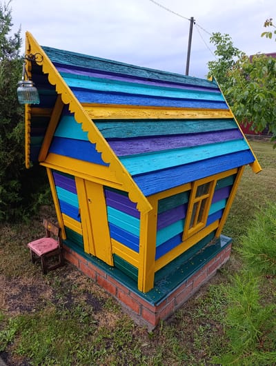 A brightly colored, miniature house sits on a brick foundation in a grassy yard. The house has a multi-colored, striped roof and walls, with alternating planks of yellow, purple, turquoise, and blue. A small yellow door and a single window with yellow trim are visible on the front. A decorative lantern hangs from the left side of the roof. Next to the house, a small, rustic wooden stool with a pink seat is placed on the grass. The surrounding environment includes green grass, some dry patches of ground, and foliage from trees and bushes. In the background, a wooden utility pole with wires and more trees can be seen. The sky is overcast. This scene is located in Белавушскі сельскі Савет, Belarus.