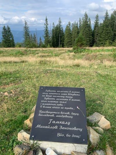The image depicts a mountainous landscape in Vorokhta, Ukraine, under a cloudy sky. The foreground features a rectangular black memorial plaque with white Ukrainian text, standing upright and supported by an arrangement of natural stones and small rocks. A small, white, cylindrical object, possibly a plastic container or bottle, lies at the base of the plaque on the left. The text on the plaque dedicates it to "Hamkal Mykhailo Zenonovych," described as a warrior, friend, teacher, and traveler, and includes a poetic verse about finding solace in nature, specifically "a piece of earth and a piece of sky." Below the name is an additional phrase, "Viyo, bo dosh!" (Viyo, because of the rain!). The plaque is positioned within a grassy field, characterized by both green and dried, light brown vegetation. Beyond the field, a dense line of tall evergreen trees forms a forest. In the far background, additional layers of mountains are visible beneath the overcast sky, indicating a high-altitude location. No people are present or actions are depicted in the scene.
