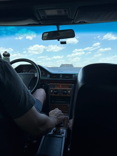 The image shows a first-person perspective from the inside of a car, looking forward through the windshield. The driver's hands are visible on the steering wheel and the gear shift. The dashboard features wood grain accents and a set of gauges, with the speedometer indicating a speed between 120 and 140. The car appears to be in motion, with the sky and clouds visible through the windshield, suggesting an outdoor environment. There is another vehicle visible in the distance ahead. The scene does not contain any elements that can be used to determine the specific city of Koshary, Ukraine, or any other geographical location.
