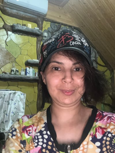 A medium shot portrait captures a woman with dark hair and a baseball cap, facing the camera. She has brown eyes and is smiling slightly. Her cap features various writings and designs in red, white, and black. She is wearing a colorful shirt with a floral and polka dot pattern. In the background, there is a shelf with bottles and jars, a textured yellow wall, and a wooden ceiling. The environment appears to be indoors, possibly a home or a workshop.
