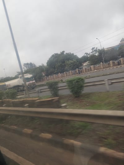 The scene depicts a multi-lane highway in Gatongora ward, Kenya, under an overcast grey sky. In the immediate foreground, a grey metal guardrail runs horizontally, with a paved shoulder and faded white and yellow road markings visible beneath it. The road ahead features a central median strip containing small, manicured green bushes and grass.
On the far lanes of the highway, a white tanker truck, partially labeled "PETROLEUM" on its tank, moves from left to right. Behind it, a yellow vehicle, possibly a bus, is partially visible. Further to the right, beyond the median, several individuals are visible walking or standing on a paved area or sidewalk, behind a decorative, light-colored stone or concrete fence with darker posts.
The background consists of dense green trees and foliage, partially obscuring a multi-story building with a light facade and a darker roof, visible on the far right. A street light pole with an extended arm stands near the building, and utility lines are discernible against the sky. The overall environment is urban or peri-urban, with ongoing vehicle and pedestrian movement.