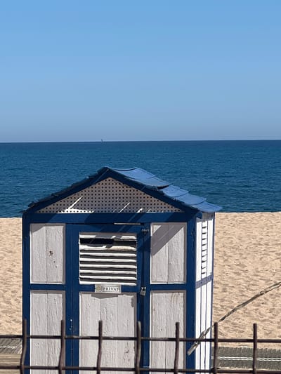 The image shows a small, weathered beach hut on the sandy coast of Sant Pol De Mar, Spain. The hut, painted in a combination of white and blue, features a lattice-style facade and a sign that reads "PRIVAT" (likely indicating private property). The hut stands with the Mediterranean Sea on its left, meeting a clear blue sky at the horizon. The hut's structural details include its wooden construction and a slatted window. The foreground of the shot features a fence or grid structure slightly out of focus. The sand on which the hut sits slopes slightly towards the sea.