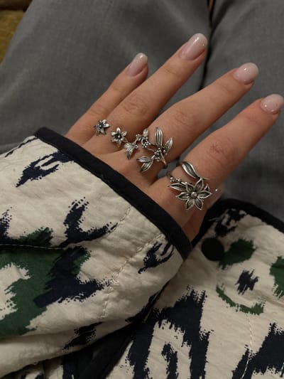 A close-up shot shows a hand adorned with two silver-colored rings. The first ring, on the index finger, features a floral design with petals extending from the center. The second ring, located on the middle finger, consists of a cascading series of three smaller flower-like elements linked together. The hand is resting against a garment with a patterned fabric that incorporates shades of cream, green, and dark blue or black, with a black trim visible along the edge. The background is a muted gray. No information suggests the image was taken in Aratashen, Armenia.