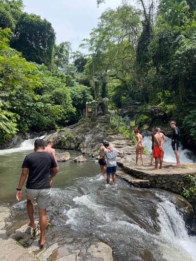 The image depicts a scene at a natural waterfall and river area in Ambengan, Indonesia, under bright daylight conditions. The environment is characterized by dense, lush tropical foliage, tall trees, and a clear river flowing over rocks, creating multiple small waterfalls and rapids.
In the center, stone steps ascend a rocky incline, leading to a small shrine or ceremonial area. This area features a dark statue, possibly a deity, situated beneath a small thatched-roof structure adorned with colorful decorations. To the right of the steps, a rustic wooden footbridge spans a section of the river.
A total of eight individuals are present in the scene, all appearing to be adults engaged in recreational activity.
On the right side, a group of five individuals stands on a stone platform adjacent to the river. This group includes one female with reddish hair, facing away from the viewer, dressed in a white two-piece swimsuit. Beside her, one male wears red swim shorts, facing left. Another male wears a black long-sleeved swim shirt and black shorts, facing away. Two other males are partially visible wading in the shallow water directly in front of the platform; one wears light blue shorts and a grey-patterned short-sleeved shirt, while the other is mostly obscured.
In the foreground and to the left, three additional males are either walking barefoot on the rocky riverbed or wading through the shallow water. The closest male, in the immediate foreground, faces away from the viewer, wearing a black t-shirt and light grey shorts. Behind him, another male, shirtless, wears light-colored shorts and walks in the water. A third male is partially visible further back in the water.
The people are either wading, standing, or walking near the water, suggesting activities related to swimming, exploring, or observing the natural surroundings. The water flows vigorously over the rocks, indicating a current.