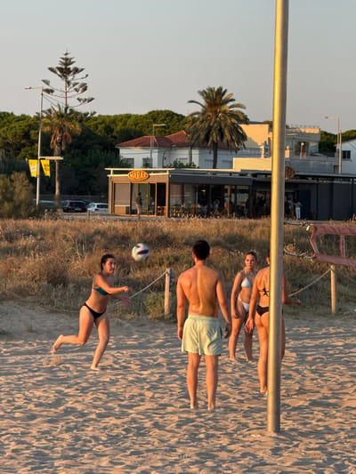 A group of young adults are playing beach volleyball on a sandy beach in Castelldefels, Spain. The scene takes place during the golden hour, with the warm sunlight casting long shadows across the sand, suggesting it is late afternoon or early evening. The sky is clear and pale, indicating fair weather.

In the foreground, a man wearing light green shorts is lying on the sand, his torso visible. Next to him, a woman in a black bikini is in the process of playing volleyball, reaching up to hit the ball. She appears focused and active. Further back, another woman, also in a bikini, is lying down on the sand, looking towards the sky. Behind her, two more individuals are partially visible, also relaxing or playing.

The background shows a building with a sign that reads "MARAE" in yellow lettering, suggesting a nearby establishment such as a restaurant or beach bar. Palm trees and other vegetation are visible behind the building, adding to the coastal atmosphere. A volleyball net is set up, confirming the activity. The overall mood seems relaxed and recreational, capturing a typical summer day at the beach.
