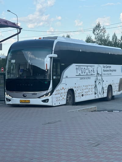 The image shows a white and black Yutong bus parked on a paved area in the city of Yarkovo, Russia. The bus has the number "1402" displayed on the front and side, and features an image of Dmitry Mendeleev and text in Russian. The text on the bus reads "ТОБОЛЬСК", "СИБИРСКИЙ ЭКСПРЕСС", and "Борт Дмитрия Ивановича Менделеева". The license plate of the bus is "AE 535 72 RUS". The background includes trees, a lamppost, a roof structure, and a cloudy sky. A red and white circular sign with a horizontal bar is visible on a fence to the left of the bus. The ground is paved with interlocking bricks.