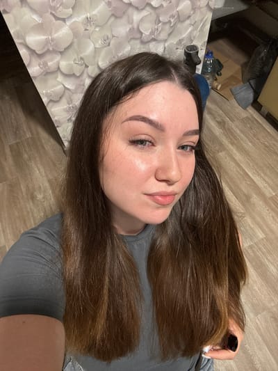 A young woman with long, brown hair is taking a selfie indoors. She is wearing a gray t-shirt. The background includes a wall with an orchid pattern, a wooden floor, and various objects on the floor, including a bottle of water, a broom, and cardboard boxes. The scene appears to be a domestic setting. The location is not specified as Shirkovo, Russia, nor can it be inferred from the image.