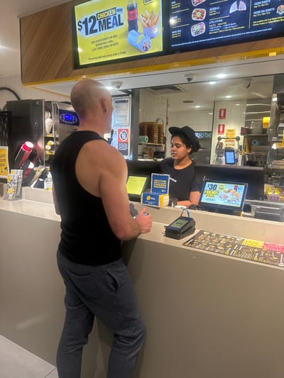 The image depicts an indoor scene at a fast-food restaurant, likely Guzman y Gomez (GYG), given the visible text. The primary subjects are two individuals: a cashier with dark curly hair, wearing a black t-shirt and a black baseball cap, and a customer with a shaved head, wearing a black tank top. The cashier is looking down at a screen, appearing to be serving the customer. The customer is facing away from the camera, looking towards the counter and holding a phone.
The setting is a brightly lit restaurant counter. Behind the cashier, there are menu boards displaying food items and prices. One menu board prominently features a "$12 CHICKEN MINI MEAL" with a Coca-Cola Zero bottle and fries. Another screen displays a "$30 FRIENDS FIESTA" promotion. Various objects are visible on the counter, including a payment terminal, napkins, and drink cups. In the background, shelves with food supplies and kitchen equipment can be seen.
The activity taking place is a transaction at the counter, with the cashier serving the customer. The overall atmosphere seems casual and typical of a fast-food establishment during operating hours. There are no strong indicators of the time of day or weather, but the indoor lighting suggests it's not dark outside. Visible text includes menu items like "CAESAR BURRITO," "QUESADILLA," and "NACHOS," as well as promotional text like "FRIENDS FIESTA" and a call to "HELP FUND WORLD-CLASS CANCER RESEARCH." The location context of North Boambee Valley, Australia, is provided externally and not directly evident from the image itself.