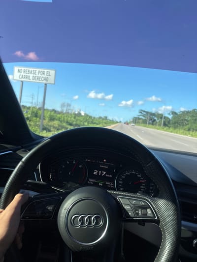 An image captured from the driver's perspective inside an Audi vehicle, identifiable by the steering wheel logo and digital dashboard. The vehicle's digital speedometer displays a speed of 217 km/h. The driver's right hand grips the left side of the black, perforated leather steering wheel. Through the windshield, a multi-lane highway is visible, stretching ahead under a bright blue sky with scattered white clouds. Dense green foliage lines both sides of the road. On the left side of the highway, a white rectangular sign with black text reads "NO REBASE POR EL CARRIL DERECHO" (DO NOT OVERTAKE ON THE RIGHT LANE). Distant vehicles are visible ahead on the road. The environment suggests a sunny day in Xcanatún, Mexico.