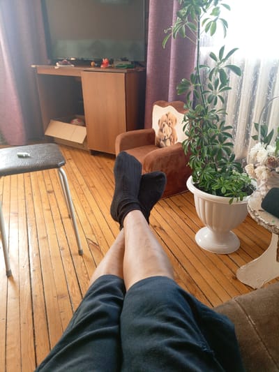 The image depicts a casual indoor scene, likely a living room, with a person's legs prominently featured in the foreground. The individual is wearing dark socks and dark shorts and is reclining, with their legs crossed.
The environment appears to be a room with light-colored wooden floorboards. To the left, a dark-colored stool with a textured seat and metal legs is visible, with a small green object resting on its surface. In the mid-ground, a brown armchair with a teddy bear motif on its back is positioned. Adjacent to the armchair is a large potted plant with green leaves, in a white decorative planter. Behind the armchair and plant, sheer white curtains are drawn, allowing bright light to filter through. A floor-to-ceiling brown curtain is visible on the left side of the image.
Further into the background, a wooden piece of furniture, possibly a desk or dresser, is situated. On its surface, several small items are scattered, including what appears to be a green wallet, a red decorative object, and a yellow sphere. A cardboard box, partially open, is placed beneath the furniture. The top of a dark television screen is visible above the furniture.
The image does not contain any specific elements that would definitively place the scene in Churapcha, Russia. The interior design elements are common and could be found in many residential settings.