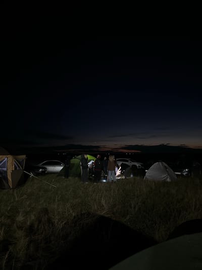 A group of people are gathered around tents at night in a grassy field. The sky is dark with a faint gradient of color near the horizon, indicating dusk or dawn. There are several vehicles parked near the tents, including a silver sedan, a white SUV, and a dark-colored car. The people appear to be engaged in conversation or setting up camp. The environment is natural, with tall grass covering the ground and a distant, indistinct landscape visible. The overall scene suggests a camping or outdoor gathering event.