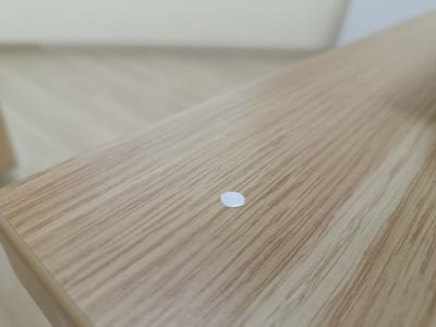 A close-up, slightly angled shot focuses on a light-colored, wood-grain textured surface, likely a table or desk. The surface has a subtle sheen, and the wood grain is characterized by thin, parallel lines of varying shades of brown and tan, running diagonally across the frame from lower left to upper right. A single, small, circular white sticker or label is positioned slightly right of the center of the frame. The background is out of focus, appearing as a pale, diffused area. The scene depicts an indoor setting. There is no identifiable information about the city of Arkadag, Turkmenistan.